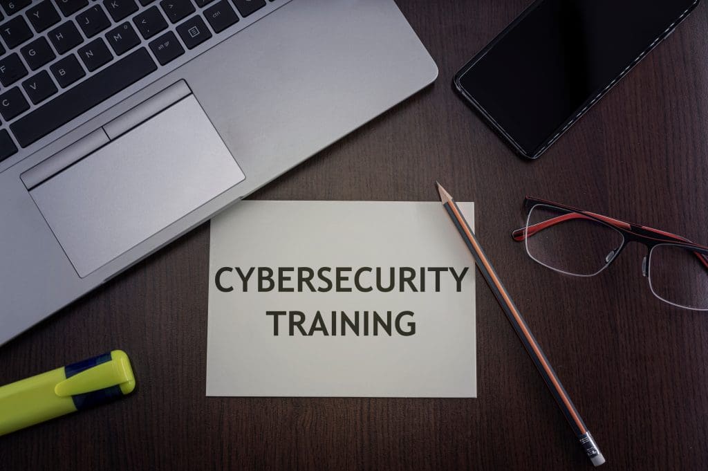 A piece of paper with the words “Cybersecurity Training” printed on it. A laptop, pencil, glasses, and a cell phone surround it.