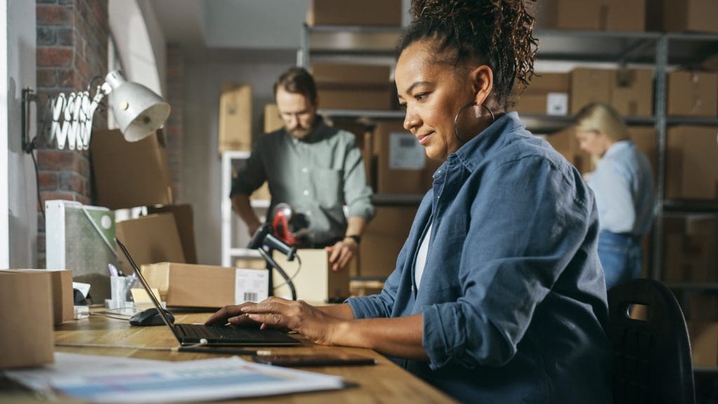 A woman sitting at a laptop while two workers pack orders in the background.