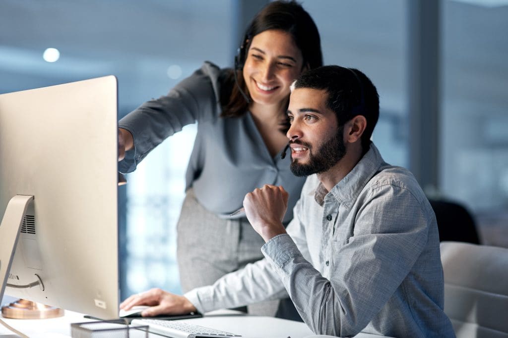 A woman smiling and pointing to a computer screen to help a co-worker.