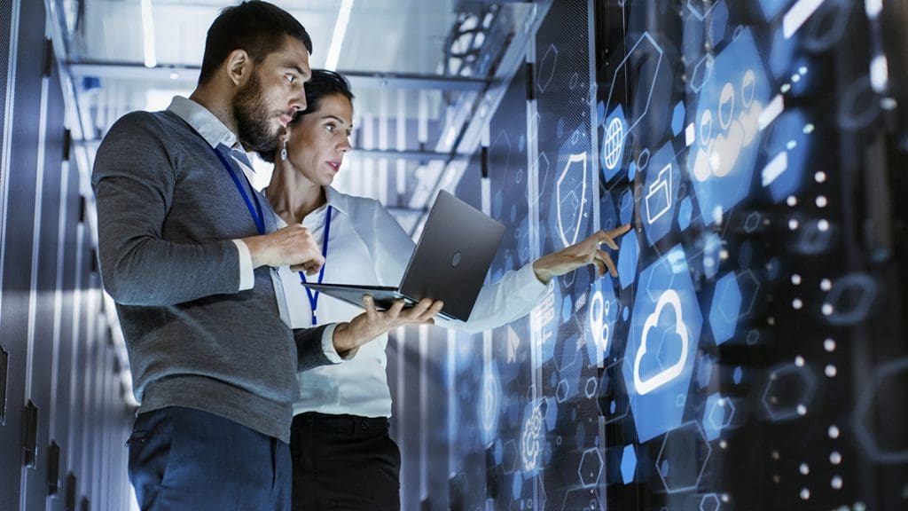 A man holding a laptop speaks with a woman pointing to a file in the server room.