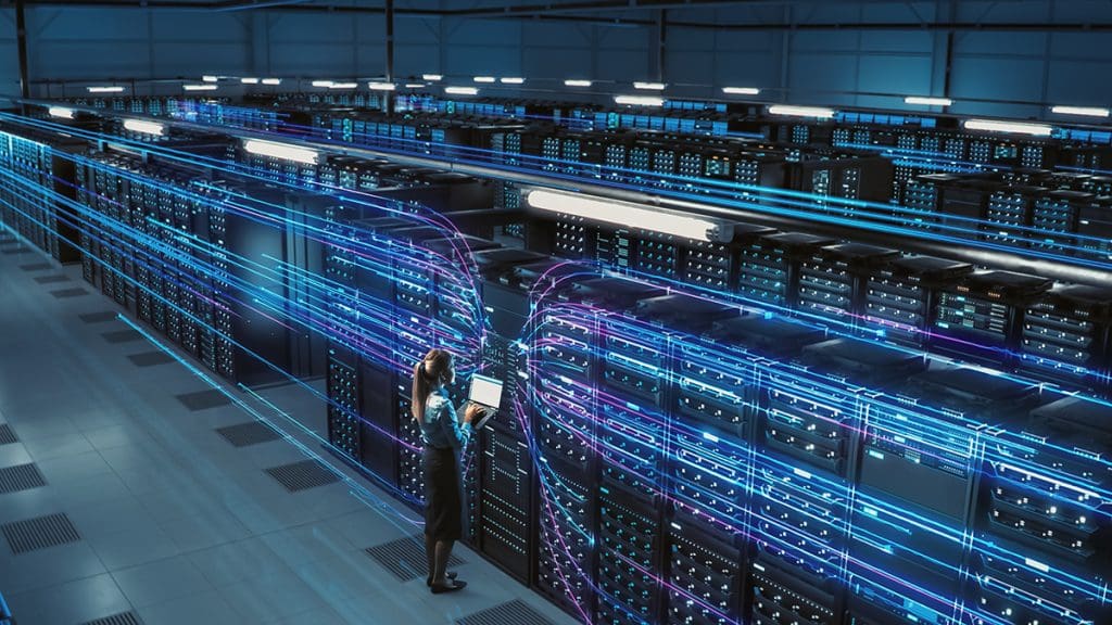 A woman stands in a server room with a laptop with neon lines leading toward her.