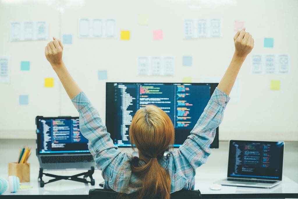 A young woman with her hands in the air in victory sitting in front of a computer.