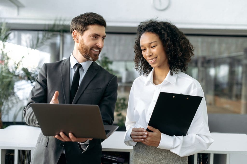 A man in a suit shows a woman in business attire some information on his laptop.