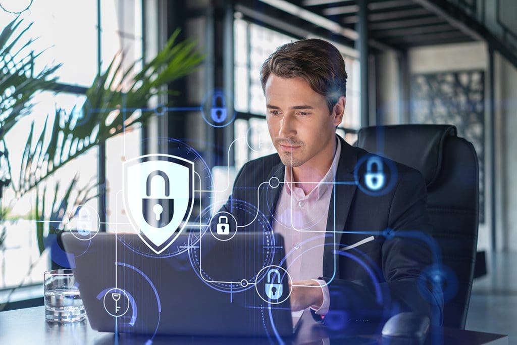 A businessman sitting at a desk in front of a laptop with cybersecurity icons overlayed on the image.