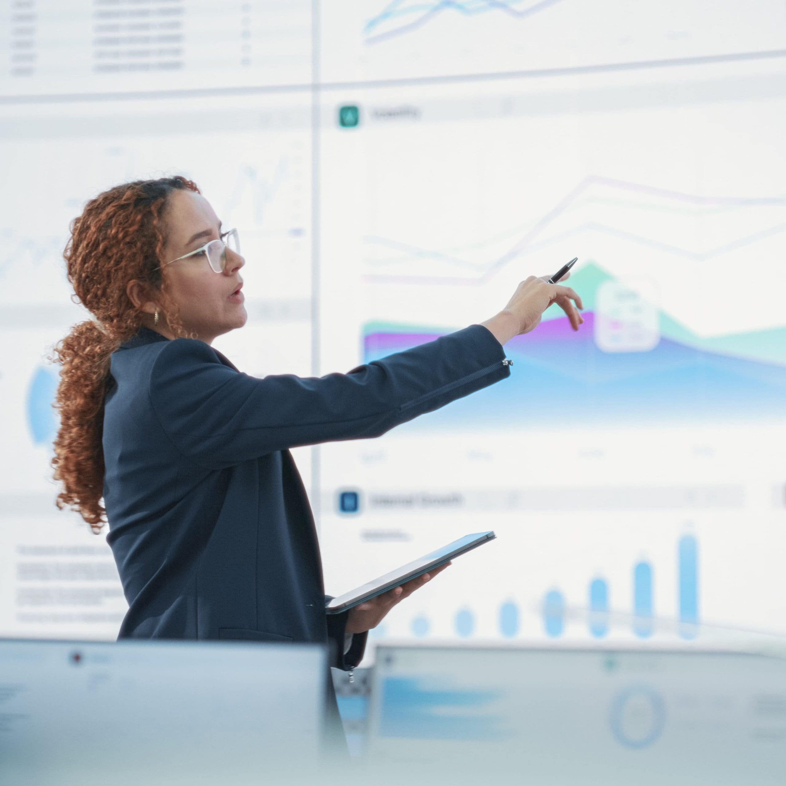  Female Senior Data Scientist Reviewing Reports Of Risk Management Department On Big Digital Screen In Monitoring Room. Diverse Consulting Company Employees Working Behind Desktop Computers.