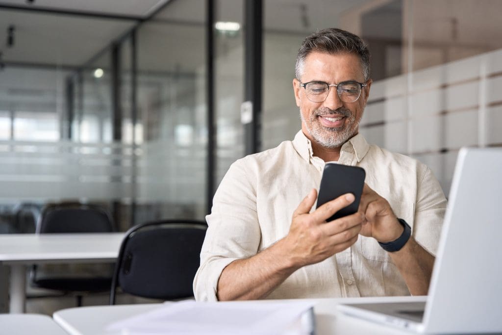 Middle aged manager ceo businessman using cell phone mobile app, laptop